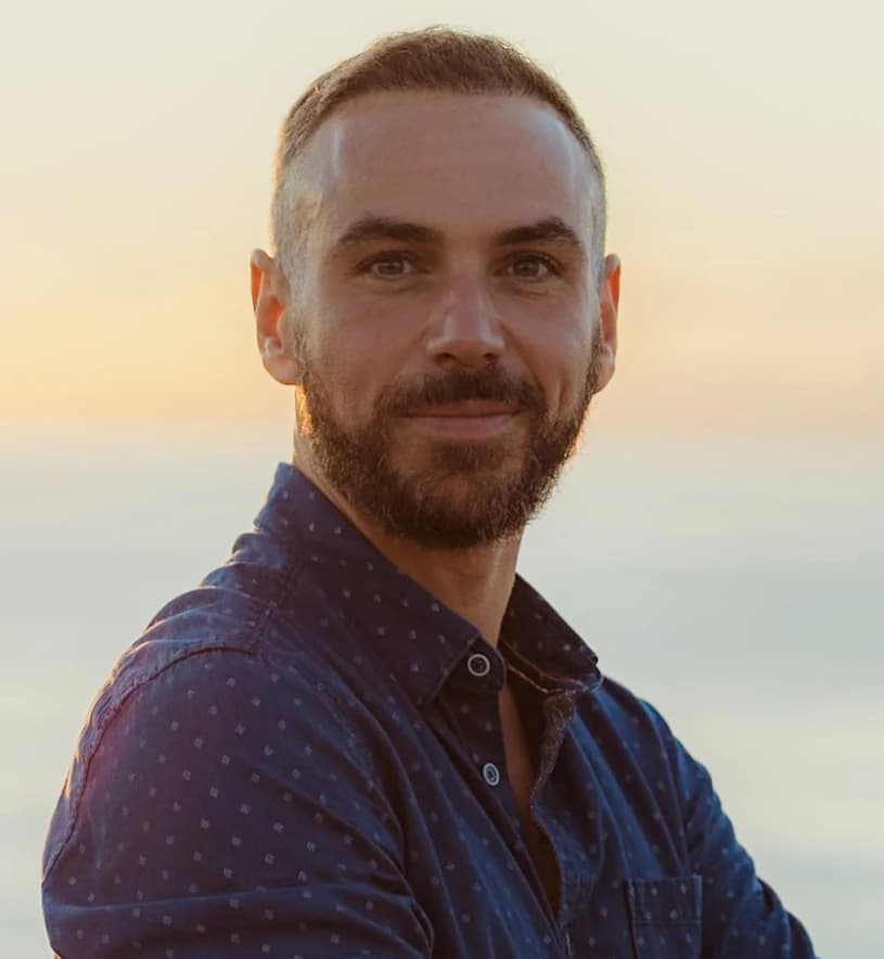 Portrait de Guillaume Churlet, souriant, en chemise bleue devant la mer au coucher du soleil